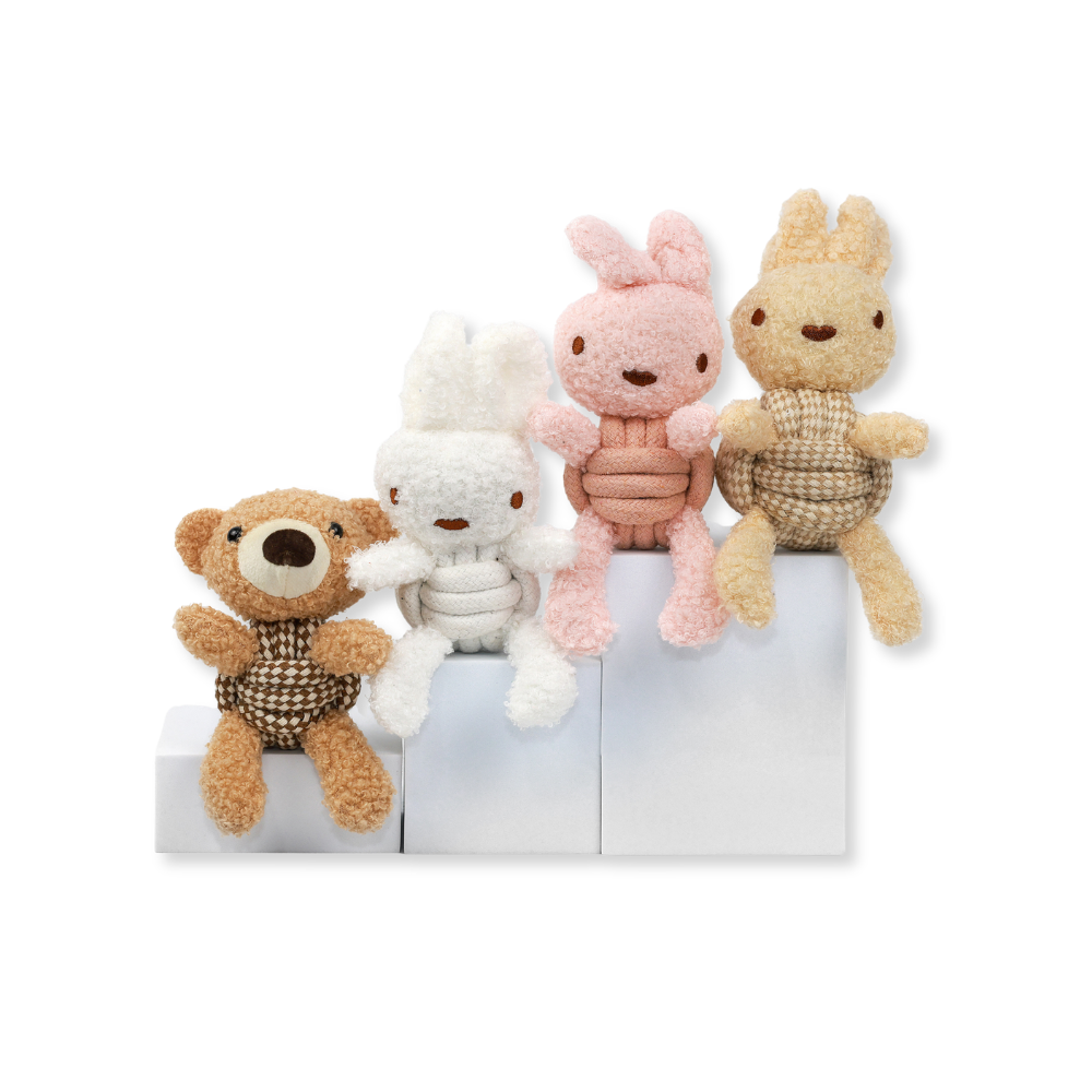 Four soft plush toys arranged on white blocks against a light gray background: a brown teddy bear, a white bunny, a pink bunny, and a tan rabbit, each wearing a knitted belly garment.